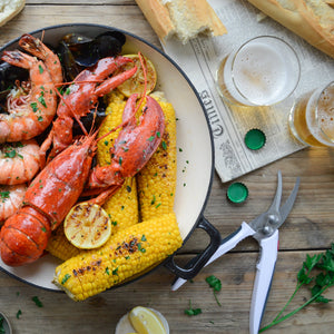 A pot of lobster, shrimp, mussels, grilled corn, and lemon sits on a rustic table with baguette and beer—plus Zyliss Shellfish Shears with stainless steel blades and a non-slip grip for easy cracking. 