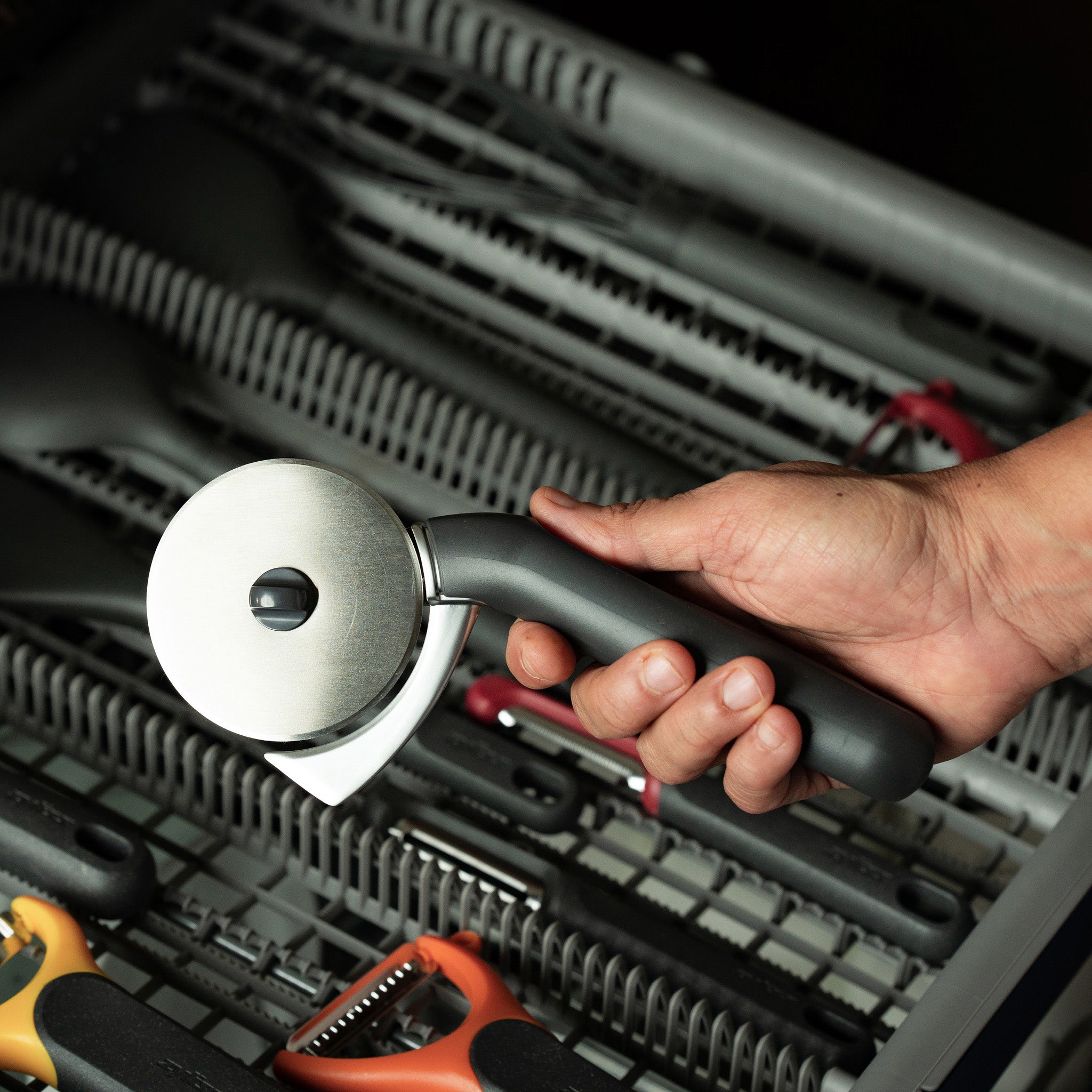 A hand holds a pizza cutter with a black handle over an open dishwasher rack filled with various kitchen utensils.