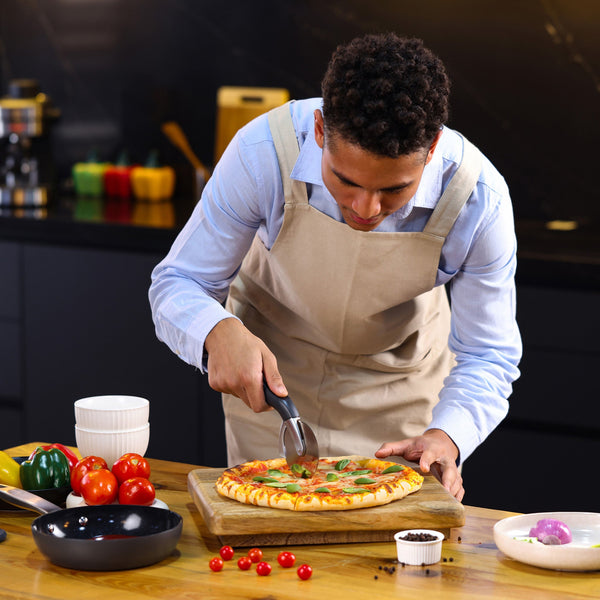 A man in a light blue shirt and beige apron uses the Zyliss Pizza & Pastry Cutter to slice a fresh pizza on a wooden board in a modern kitchen with vegetables and bowls on the counter. 