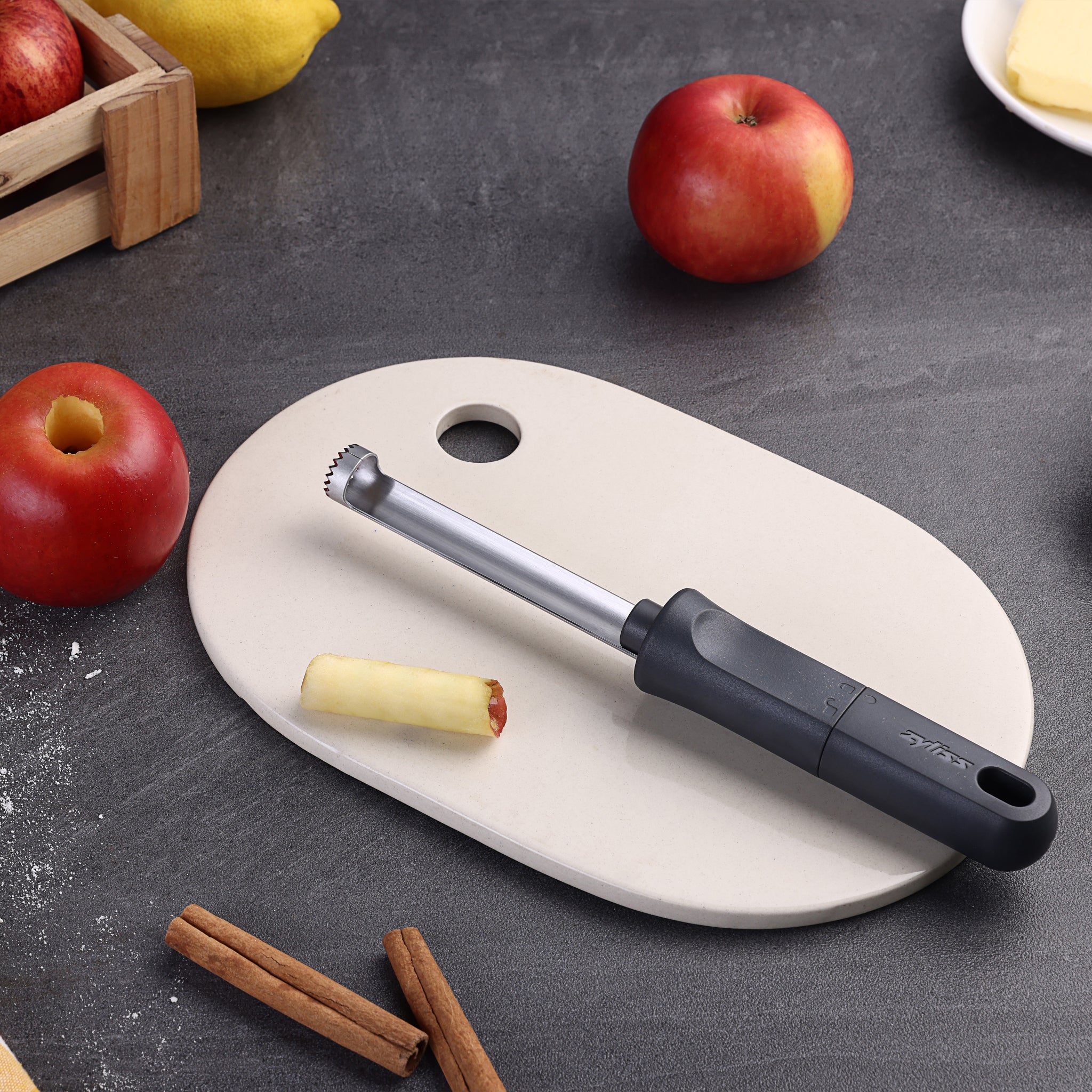 The Zyliss Apple Corer with an eco-friendly handle sits on a white cutting board next to a cored apple. Whole apples, cinnamon sticks, lemons, and a plate surround it on the dark countertop.