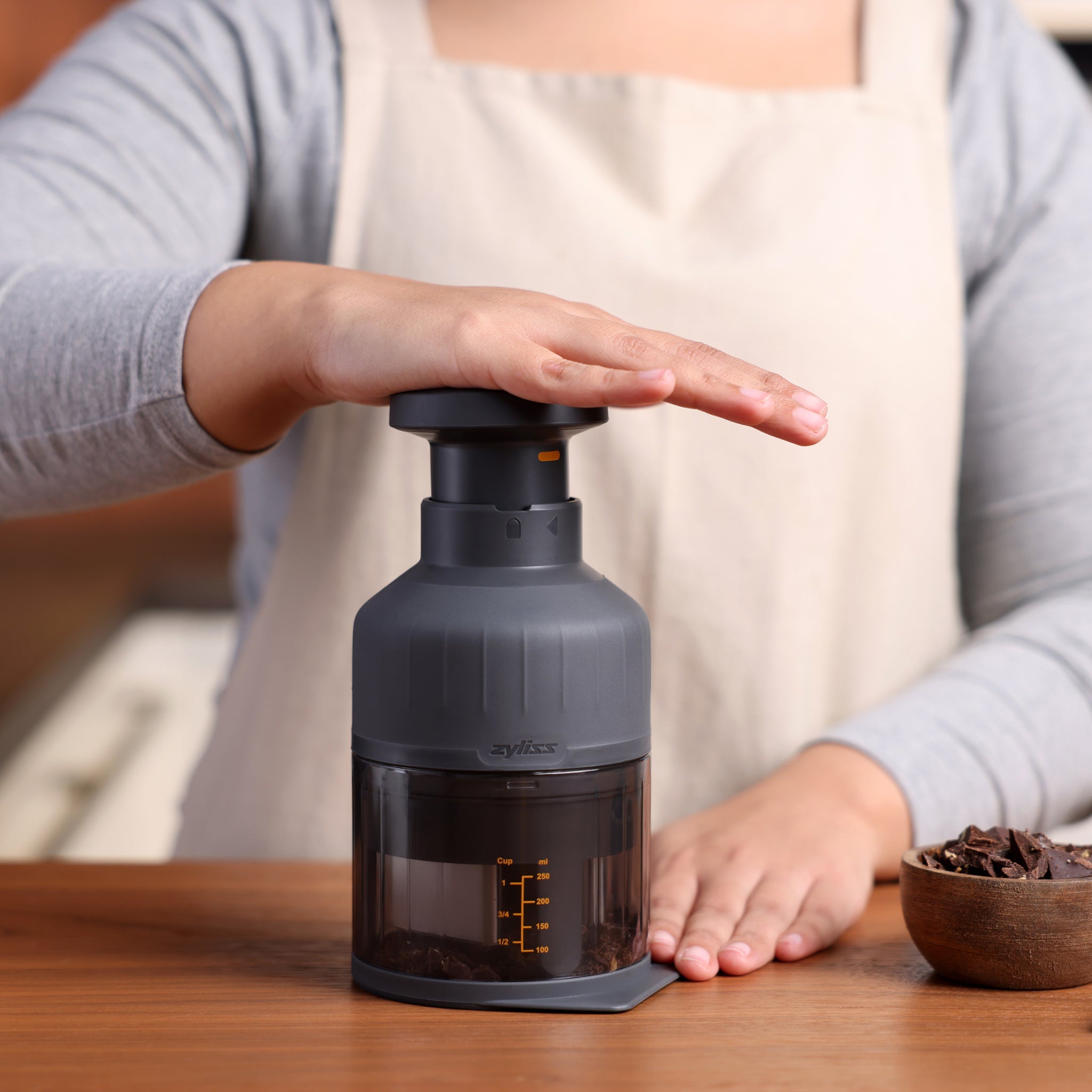 A person wearing a beige apron uses a dark gray manual food chopper on a wooden kitchen counter, with a bowl of ingredients nearby.