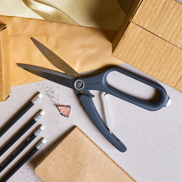 A pair of Zyliss All Purpose Shears, three black pencils with erasers, a pencil shaving, cardboard boxes, and a gold ribbon are arranged on a light-colored surface.