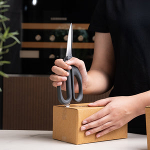 A person in a black shirt uses Zyliss Household Scissors to cut open a cardboard box on a white surface.