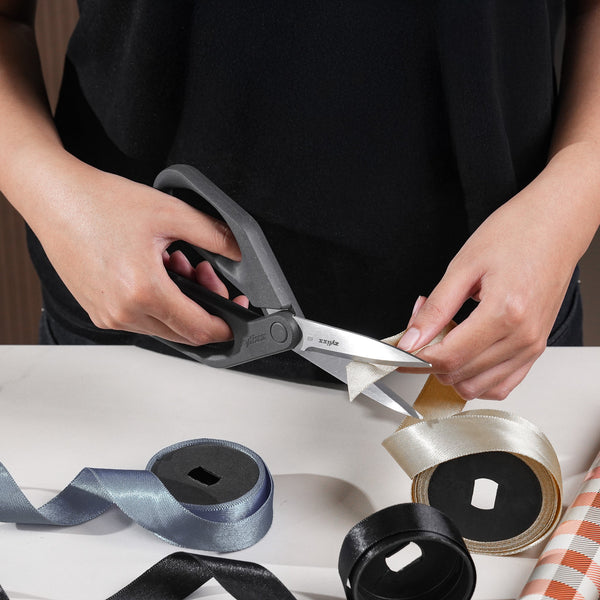 A person uses Zyliss Household Scissors with stainless steel blades to cut ivory ribbon on a table, surrounded by silver, black, and ivory rolls of ribbon.