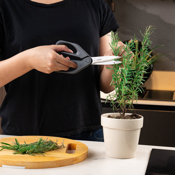 A person in a black shirt uses Zyliss Household Scissors to trim a rosemary plant in a white pot. Fresh rosemary sprigs are arranged on a wooden cutting board on a white countertop.