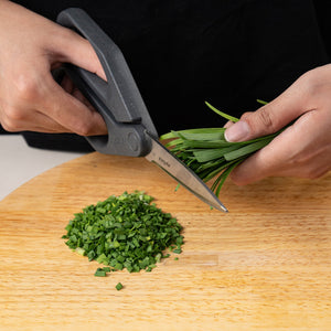 A person uses Zyliss Household Scissors with stainless steel blades to cut green herbs onto a round wooden cutting board, where finely chopped herbs are already present.