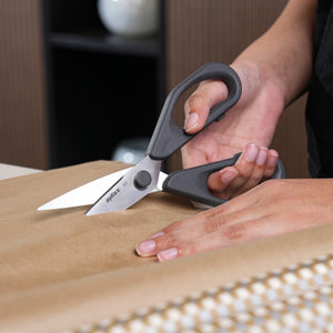 A person uses Zyliss Household Scissors Basic, featuring black handles and stainless steel blades, to cut brown paper on a flat surface, with one hand holding the scissors and the other securing the paper.
