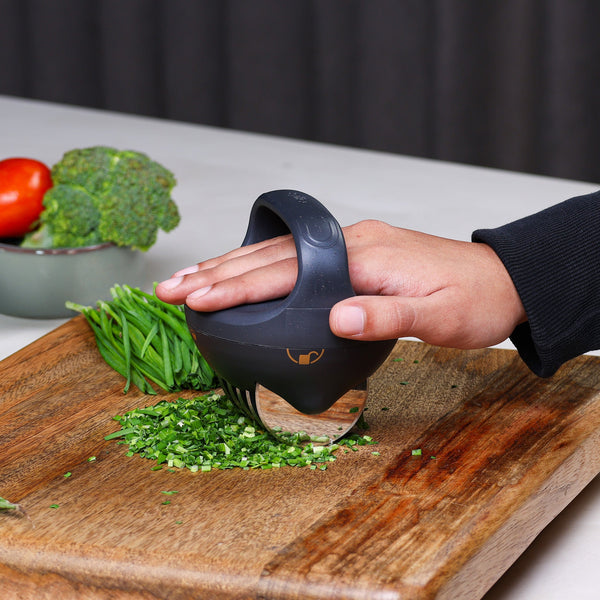 A hand uses the Zyliss Fast Cut Herb Tool, featuring stainless steel cutting wheels, to dice green herbs on a wooden cutting board. Fresh tomatoes and broccoli are visible in a bowl in the background.
