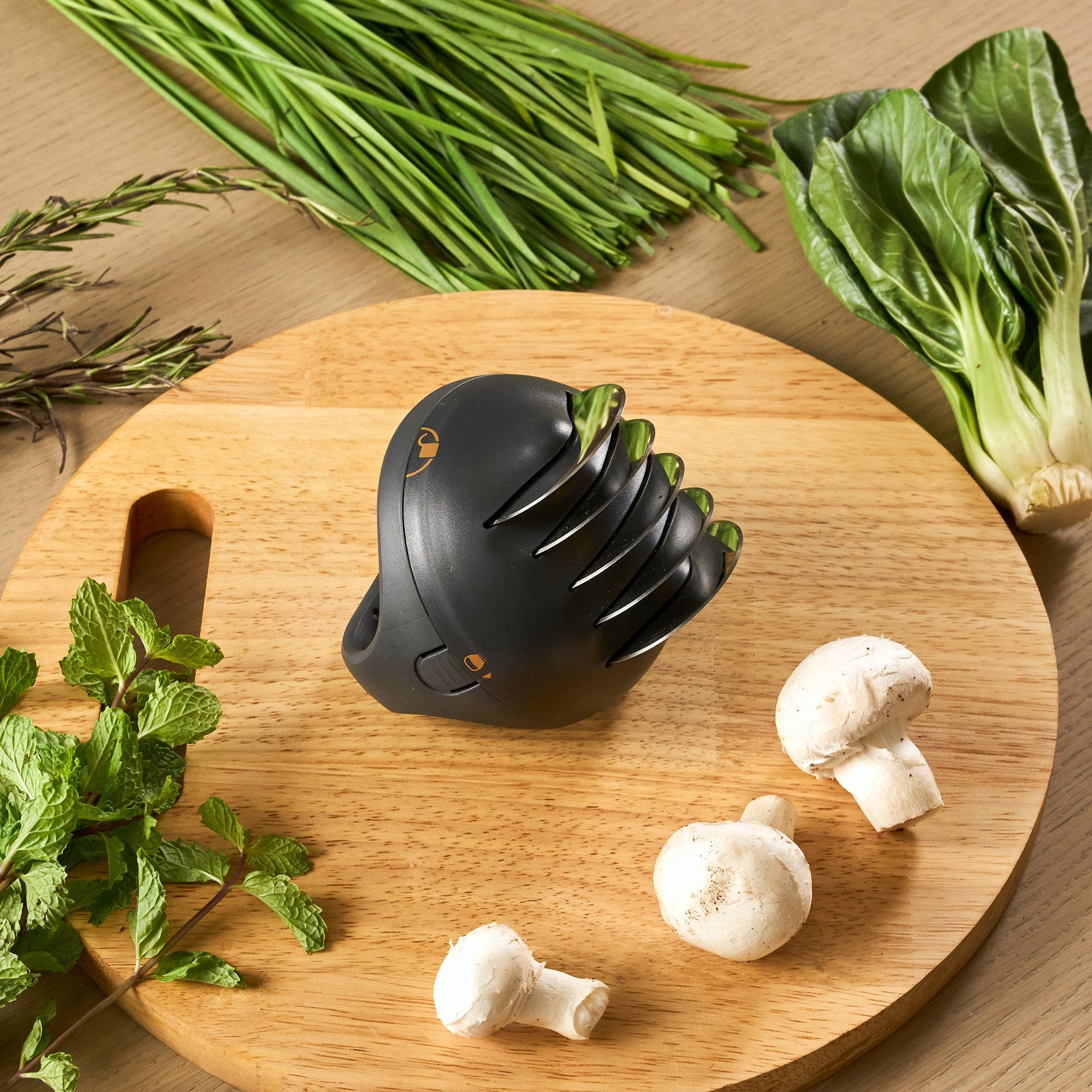 The Zyliss Fast Cut Herb Tool, featuring stainless steel cutting wheels, sits on a round wooden cutting board beside fresh herbs, leafy greens, and three white mushrooms.