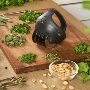 The Zyliss Fast Cut Herb Tool, featuring stainless steel blades, rests on a wooden board amid chopped herbs, fresh leaves, a bowl of peanuts, and a metal herb stripper in a cozy kitchen setting.