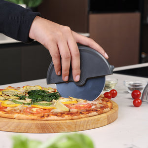 A hand slices a vegetable pizza on a wooden board using the Zyliss Pizza Wheel. Cherry tomatoes and small ingredient bowls sit on the countertop nearby.