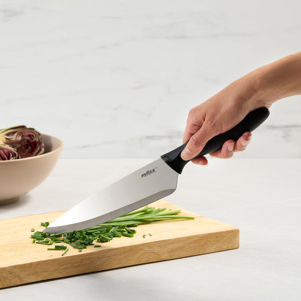 A hand uses a Zyliss 6 Piece Knife Set to chop green herbs on a wooden board. Behind, a beige bowl with artichokes rests on a white surface, showcasing essential kitchen prep tools. 