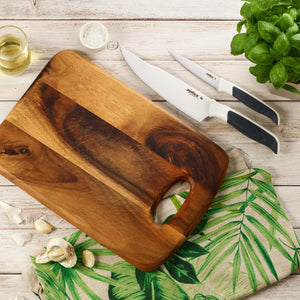 A wooden cutting board, the Zyliss Comfort Serrated Paring Knife 10.5cm/4¼, garlic cloves, a bowl of salt, a glass of oil, fresh basil, and a green leafy-patterned cloth are arranged on a white wooden surface. 