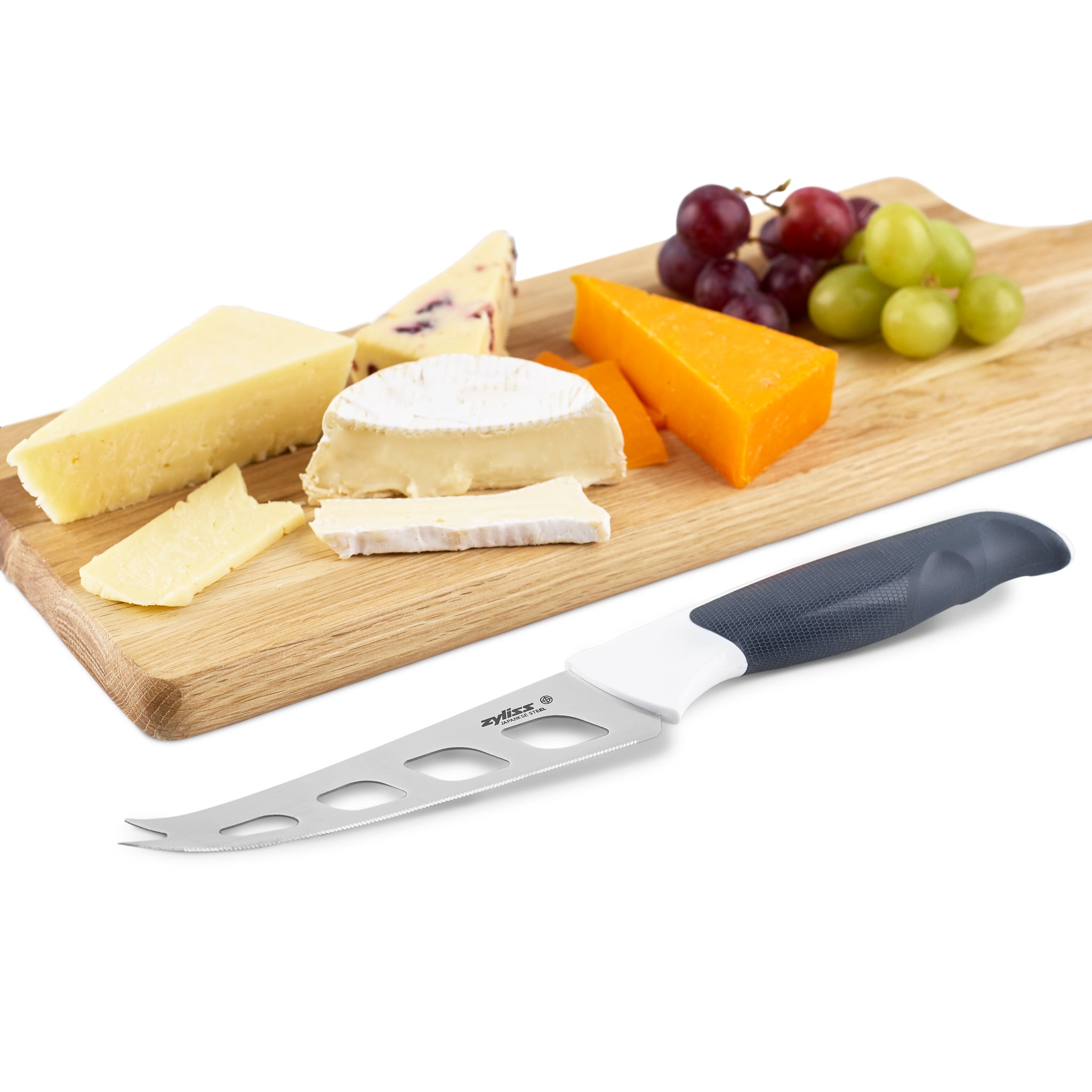 The Zyliss Comfort Cheese Knife 12cm, featuring a black and white ergonomic handle and sharp stainless steel blade, lies before a wooden board with assorted cheeses and red and green grape clusters.