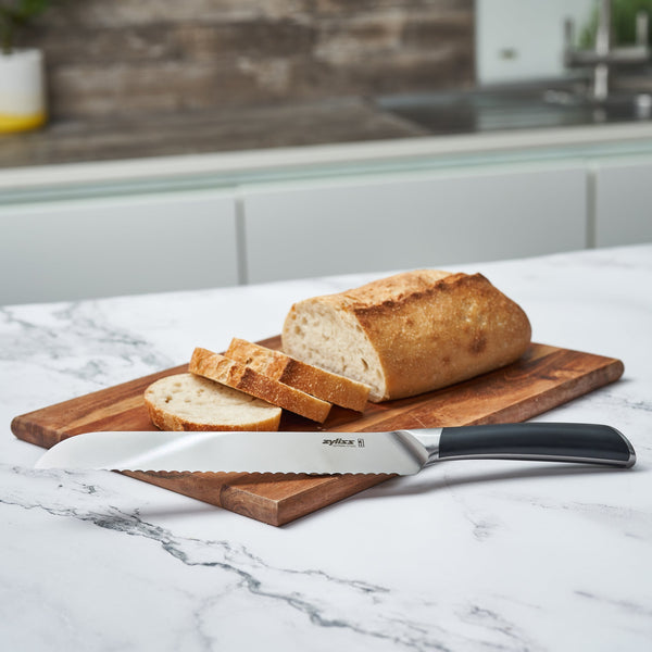 A sliced loaf of bread rests on a wooden board with the Zyliss Comfort Pro Bread Knife 20cm/8, featuring an ergonomic handle and German stainless steel blade, displayed on a marble countertop in a modern kitchen. 