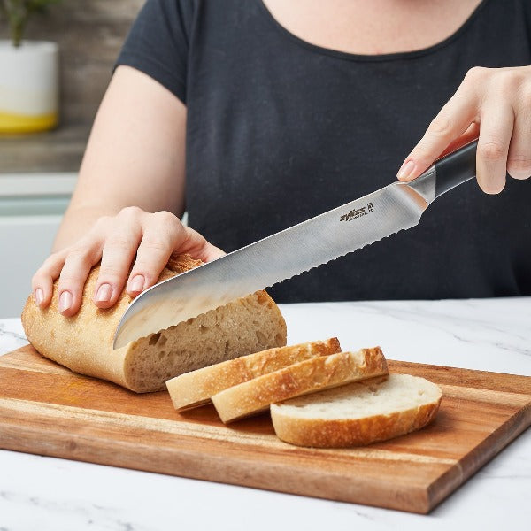 A person slices a loaf of bread with a large serrated knife on a wooden cutting board. Three slices of bread are already cut and lying in front of the loaf.
