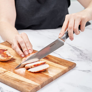 A person slices cooked chicken breast on a wooden cutting board using the Zyliss Comfort Pro Carving Knife 20cm / 8, which features an ergonomic handle, while holding the chicken steady with one hand on a white marble countertop. 