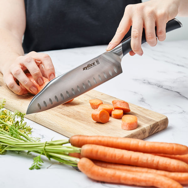 A person slices carrots on a wooden cutting board using a Zyliss Comfort Pro Santoku Knife 18cm / 7 with an ergonomic handle, while whole carrots with green tops sit beside the board on a marble countertop. 