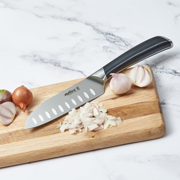 The Zyliss Comfort Pro Mini Santoku Knife 13cm rests on a wooden cutting board next to chopped shallots, whole garlic bulbs, and shallots on a white marble surface. 