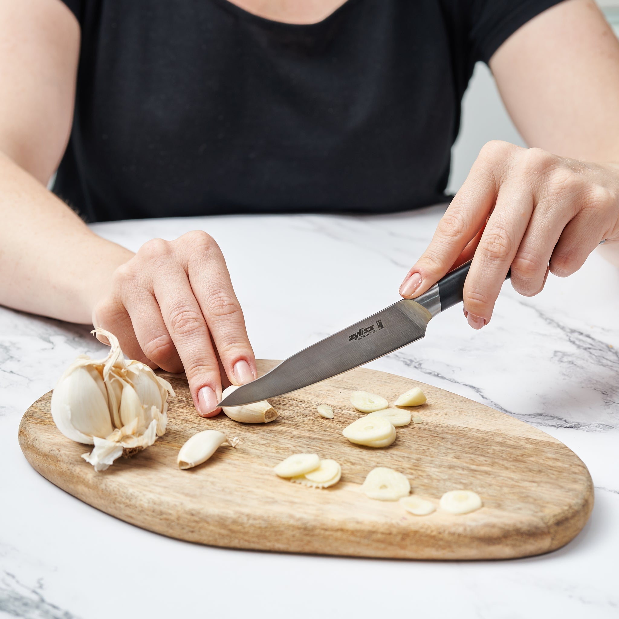 A person in a black shirt slices garlic cloves on a wooden cutting board using the Zyliss Comfort Pro 3 Piece Paring Knife & Utility Knife Set. A whole garlic bulb rests on the board above a white marble surface.