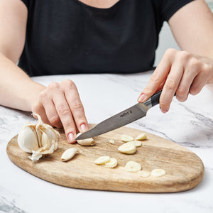 A person in a black shirt slices garlic cloves on a wooden cutting board using the Zyliss Comfort Pro 3 Piece Paring Knife & Utility Knife Set. A whole garlic bulb rests on the board above a white marble surface. 