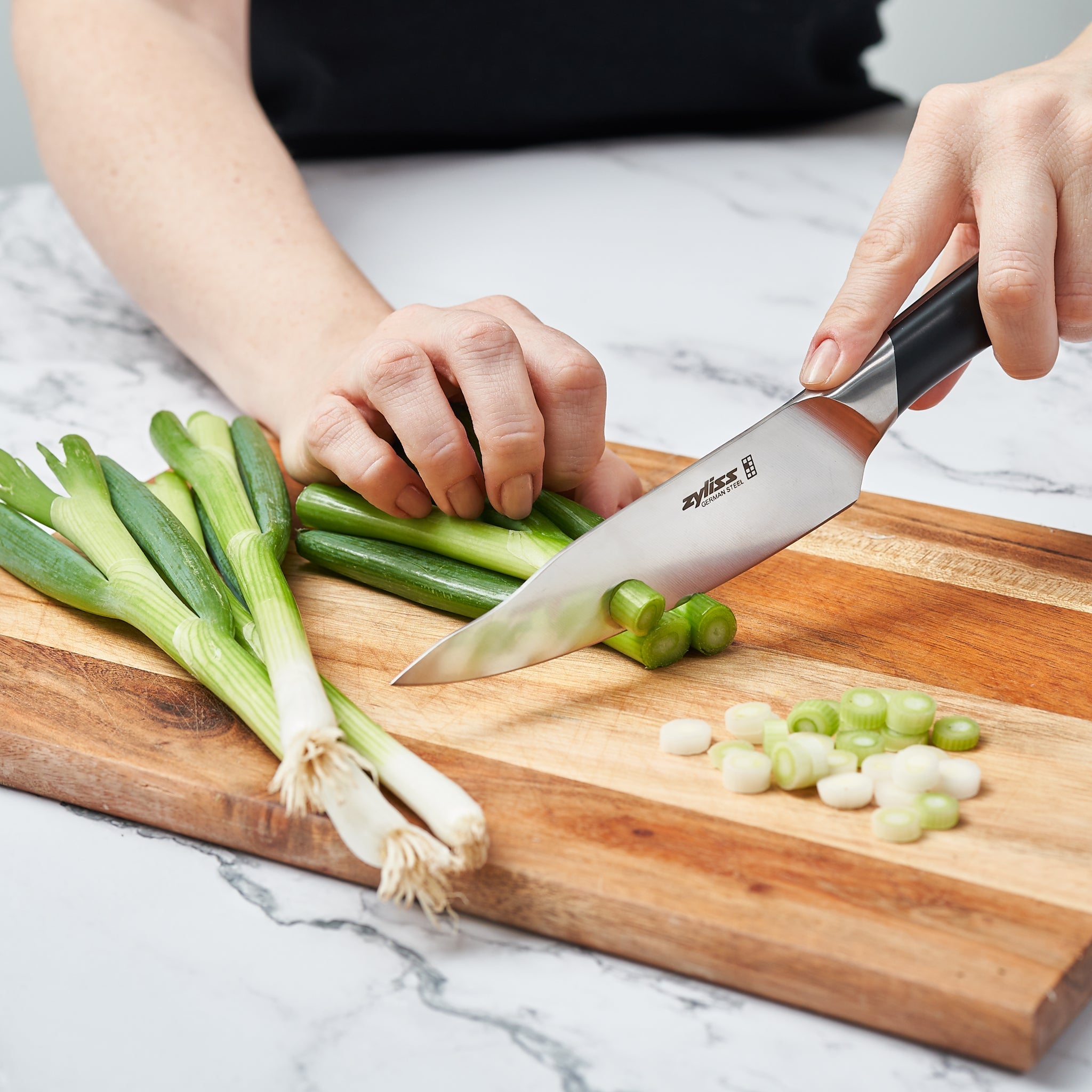 Using the Zyliss Comfort Pro Utility Knife 14cm / 5½, a person slices green onions on a wooden cutting board, with whole onions and neatly chopped pieces arranged beside the ergonomic-handled knife.