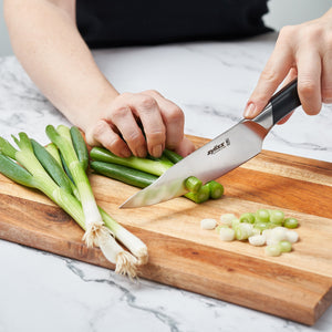 Using the Zyliss Comfort Pro Utility Knife 14cm / 5½, a person slices green onions on a wooden cutting board, with whole onions and neatly chopped pieces arranged beside the ergonomic-handled knife. 