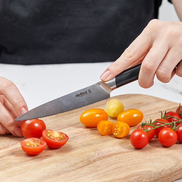 A person slices cherry tomatoes with the Zyliss Comfort Pro Serrated Paring Knife (11cm/4½, black handle, German stainless steel) on a wooden board, with whole and halved red and yellow tomatoes arranged nearby. 
