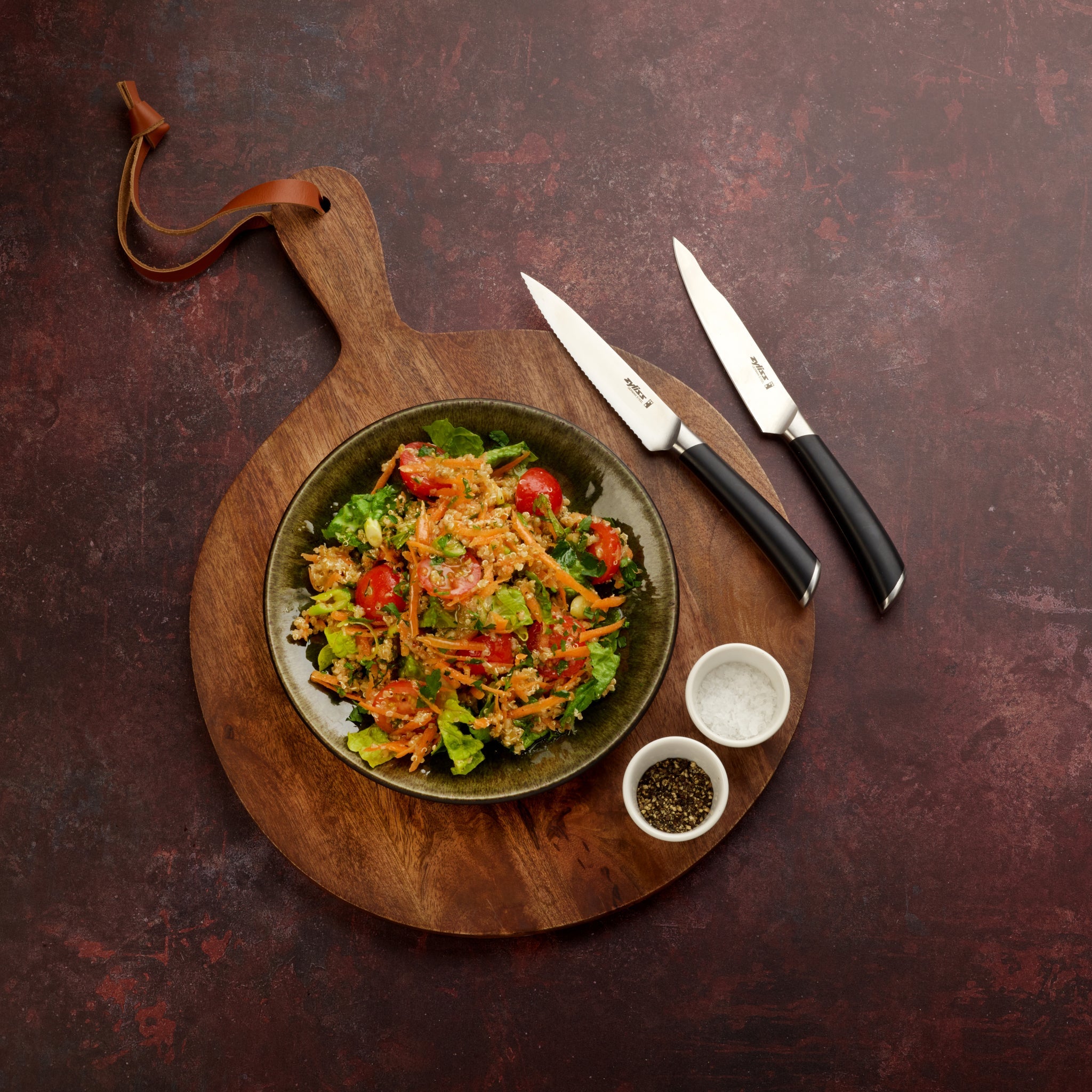 A bowl of fresh salad with greens, tomatoes, and other vegetables sits on a wooden board with two knives and small bowls of salt and pepper on a dark textured surface.
