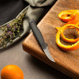 A Zyliss Peeling Knife 6cm / 2Â½ with an ergonomic handle sits on a wooden cutting board beside orange peels, while an unpeeled orange and dried herbs are nearby on a gray surface. 