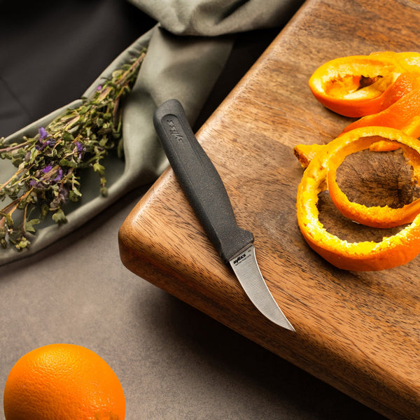 A Zyliss Peeling Knife 6cm / 2Â½ with an ergonomic handle sits on a wooden cutting board beside orange peels, while an unpeeled orange and dried herbs are nearby on a gray surface. 