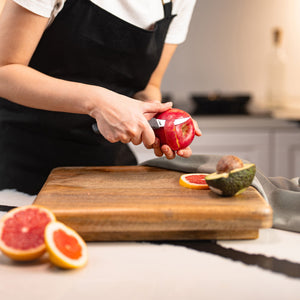 Wearing a black apron, someone peels a pomegranate with the Zyliss Peeling Knife 6cm / 2Â½ over a wooden cutting board in the kitchen, next to sliced grapefruit and avocado. 
