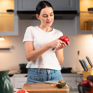 A woman in a white tee and jeans peels a red apple with the Zyliss Peeling Knife 6cm/2Â½, which has an ergonomic handle, in a modern kitchen beside a wooden cutting board filled with assorted fruits. 