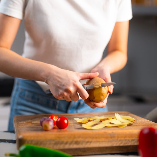 A person in a white shirt and jeans peels a potato with the Zyliss Paring Knife 9cm / 3Â½ (eco-friendly handle) over a wooden cutting board covered in potato peels, cherry tomatoes, and shallots. 