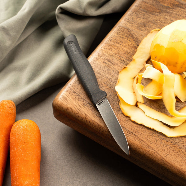 The Zyliss Paring Knife 9cm / 3Â½ with an eco-friendly handle rests on a wooden cutting board next to a peeled potato, potato skins, two unpeeled carrots, and a green cloth. 