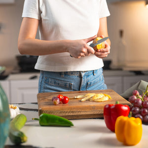 Wearing a white shirt and jeans, a person uses the Zyliss Paring Knife 9cm / 3Â½ with an eco-friendly handle to slice a potato on a wooden cutting board in a kitchen amid fresh vegetables and grapes. 