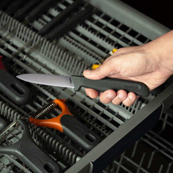 A hand holds a paring knife with a black handle above an open dishwasher rack filled with various kitchen utensils and peelers.
