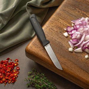A Zyliss Vegetable Knife 9cm sits on a wooden cutting board next to chopped red onions, with sliced red chili peppers and fresh thyme close by. A green cloth in the background adds a pop of color. 