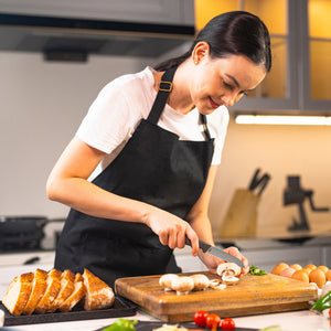 A woman in a black apron slices mushrooms with the Zyliss Vegetable Knife 9cm / 3Â½ on a wooden cutting board in a modern kitchen, surrounded by bread, eggs, and vegetables. 