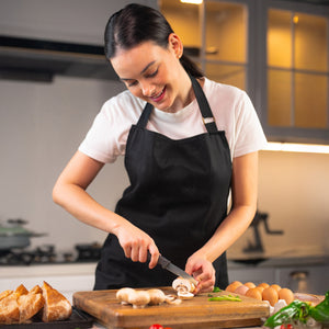 A woman in a black apron and white shirt smiles as she uses the Zyliss Vegetable Knife 9cm / 3Â½ with an eco-friendly handle to precisely slice mushrooms on a wooden board in her kitchen, surrounded by eggs, herbs, and bread. 