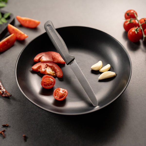 A black plate holds sliced tomatoes, three garlic cloves, and a Zyliss Tomato Knife Serrated 11cm / 4½. Nearby, whole cherry tomatoes and tomato wedges sit on a dark surface. 