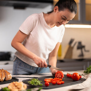 In a modern kitchen, a woman in a white t-shirt slices a tomato on a cutting board using the Zyliss Tomato Knife Serrated 11cm / 4½ with an eco-friendly handle. Sliced bread, arugula, and assorted vegetables are on the nearby countertop. 