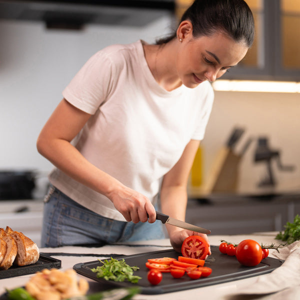 In a modern kitchen, a woman in a white t-shirt slices a tomato on a cutting board using the Zyliss Tomato Knife Serrated 11cm / 4½ with an eco-friendly handle. Sliced bread, arugula, and assorted vegetables are on the nearby countertop. 