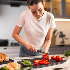 A woman in a white t-shirt slices tomatoes on a black cutting board in a modern kitchen using the Zyliss Tomato Knife Serrated 11cm / 4½, which has an eco-friendly handle. Fresh vegetables and bread are spread out on the counter around her. 