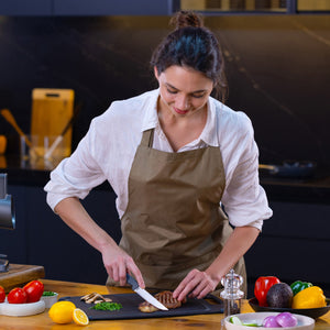 In a modern kitchen, a woman in a beige apron slices grilled chicken on a cutting board with the Zyliss Steak Knife Serrated 11cm / 4½, surrounded by fresh tomatoes, peppers, lemon, avocado, and herbs. 