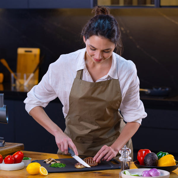 In a modern kitchen, a woman in a beige apron slices grilled chicken on a cutting board with the Zyliss Steak Knife Serrated 11cm / 4½, surrounded by fresh tomatoes, peppers, lemon, avocado, and herbs. 
