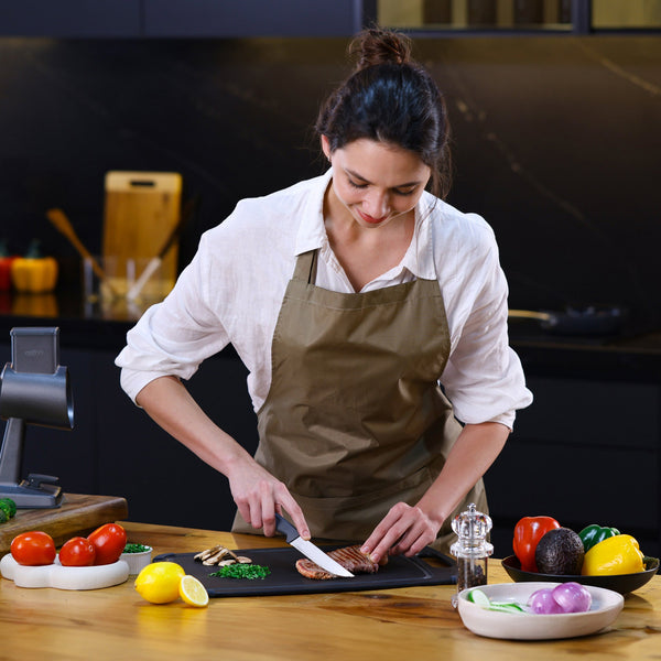 In a modern kitchen, a woman in a brown apron slices grilled chicken with the Zyliss Steak Knife Serrated 11cm / 4½ on a cutting board, surrounded by fresh vegetables like tomatoes, avocado, onion, and bell peppers. 