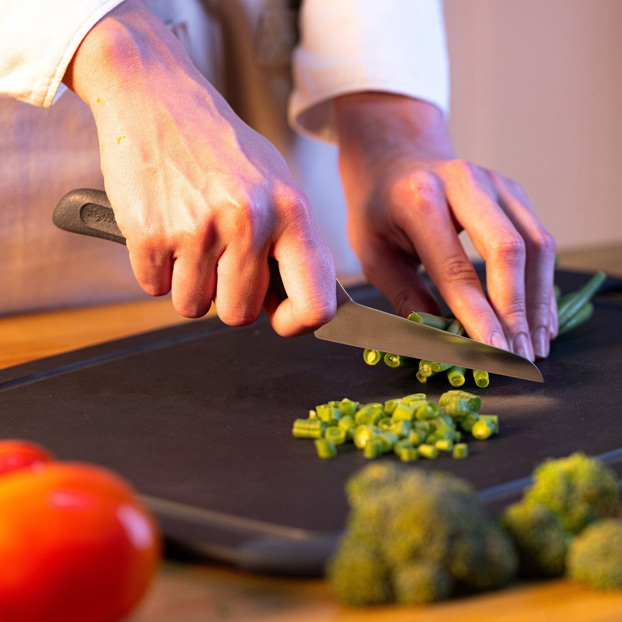 A person slicing green beans on a black cutting board with a knife, surrounded by fresh vegetables like tomatoes and broccoli.