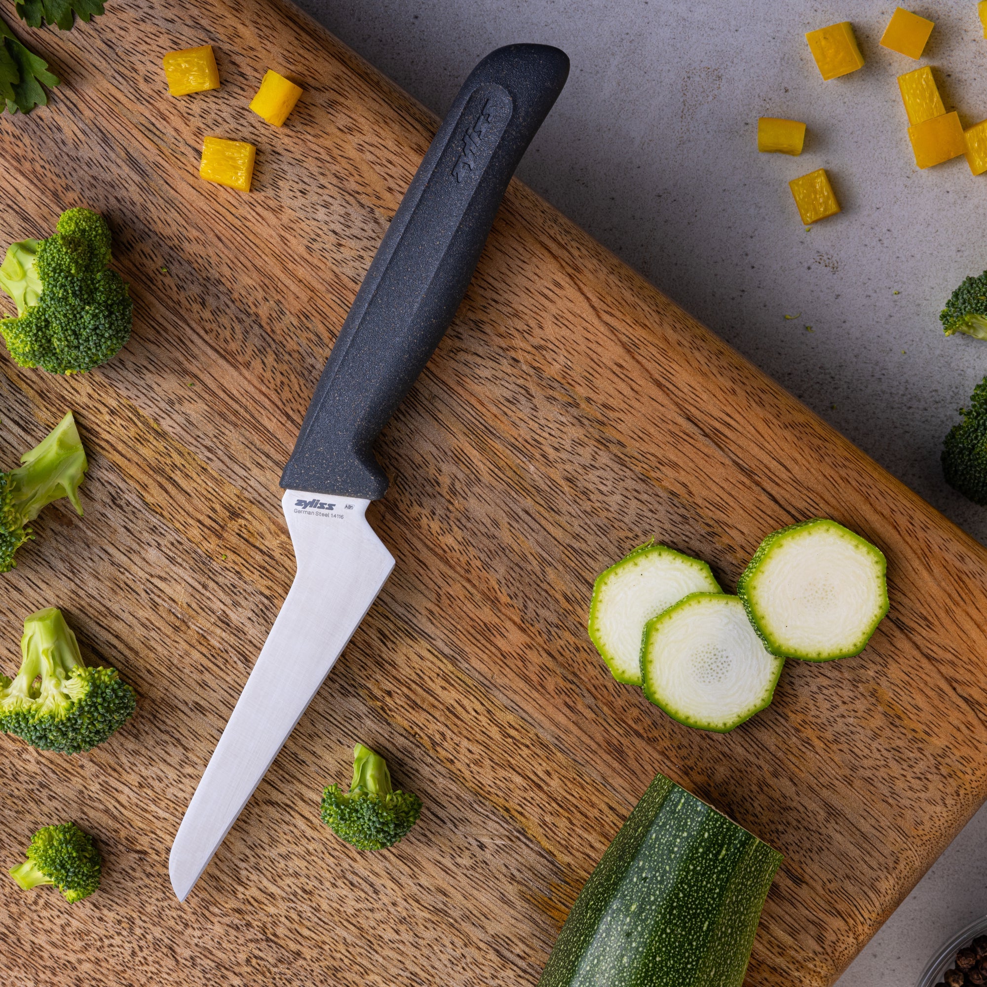 A knife with a black handle rests on a wooden cutting board surrounded by broccoli florets, yellow pepper cubes, and sliced zucchini on a gray countertop.