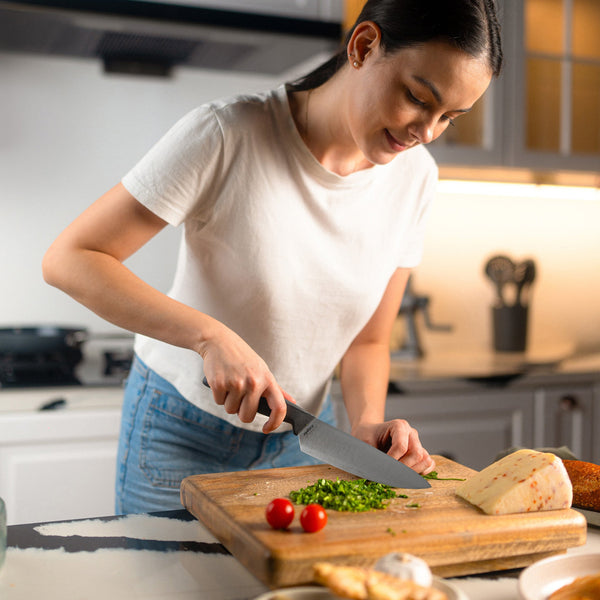 #15cm/6"
A woman in a white t-shirt smiles as she chops green herbs with the Zyliss Chef Knife 15cm/6, featuring an ergonomic handle, on a wooden cutting board in a modern kitchen. Cheese and tomatoes are also on the board.